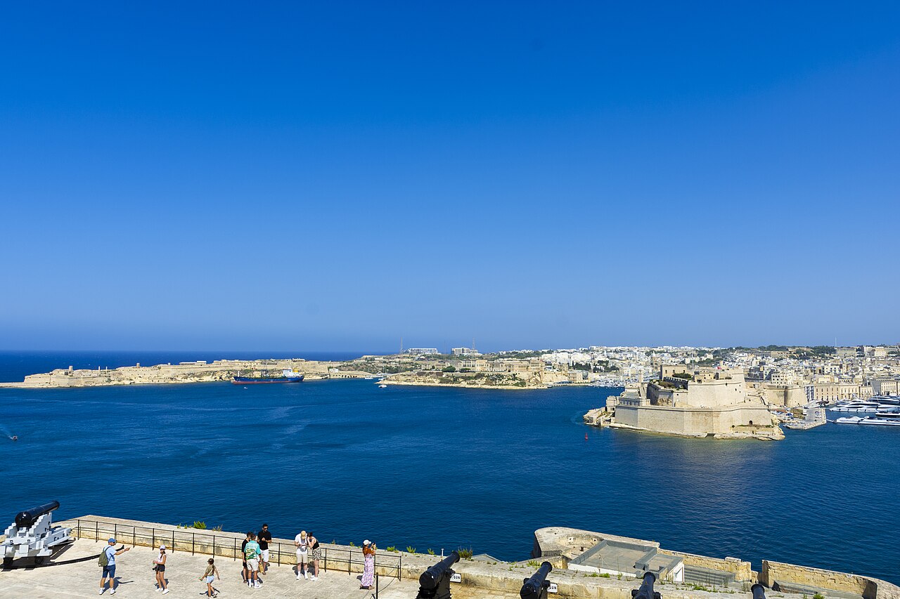 Deep blue water fills a wide harbour edged by pale stone fortifications under a cloudless sky.