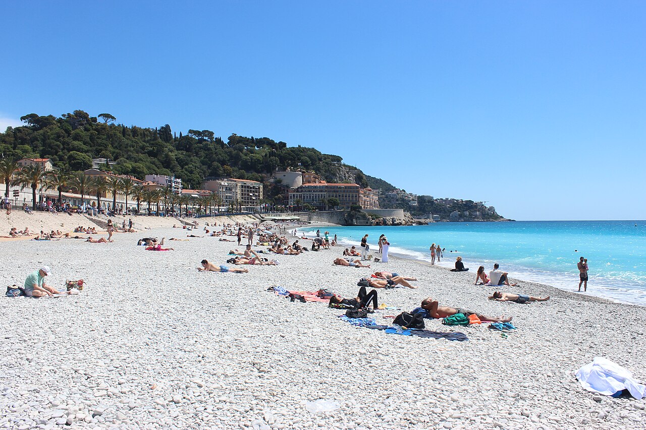 A pebble beach with scattered sunbathers curves along turquoise water towards a wooded headland.