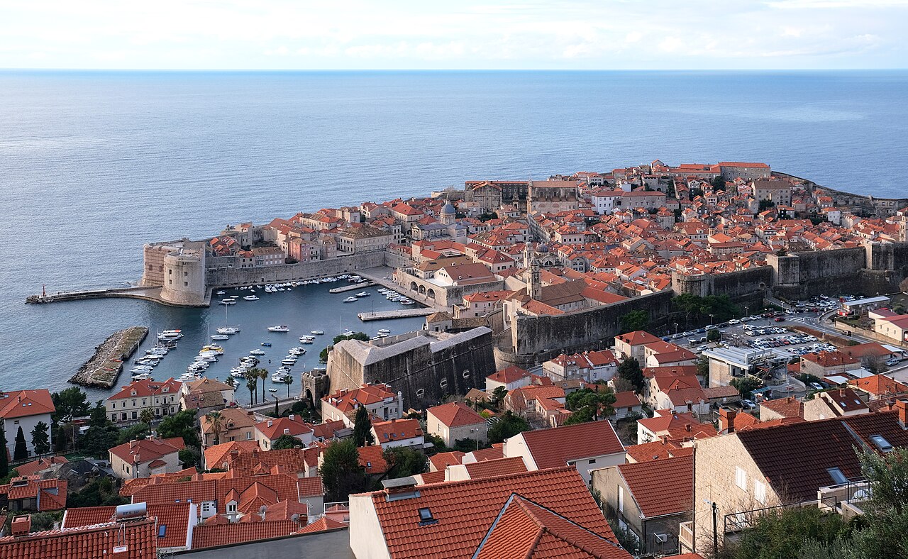 A walled town of orange-tiled roofs juts into the sea beside a small harbour full of boats.