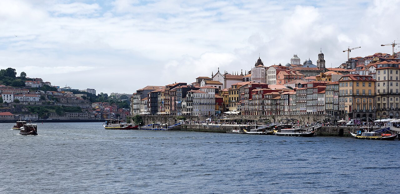 Tall, narrow, many-coloured houses line a riverbank beneath a cloudy sky, seen across a wide stretch of water.