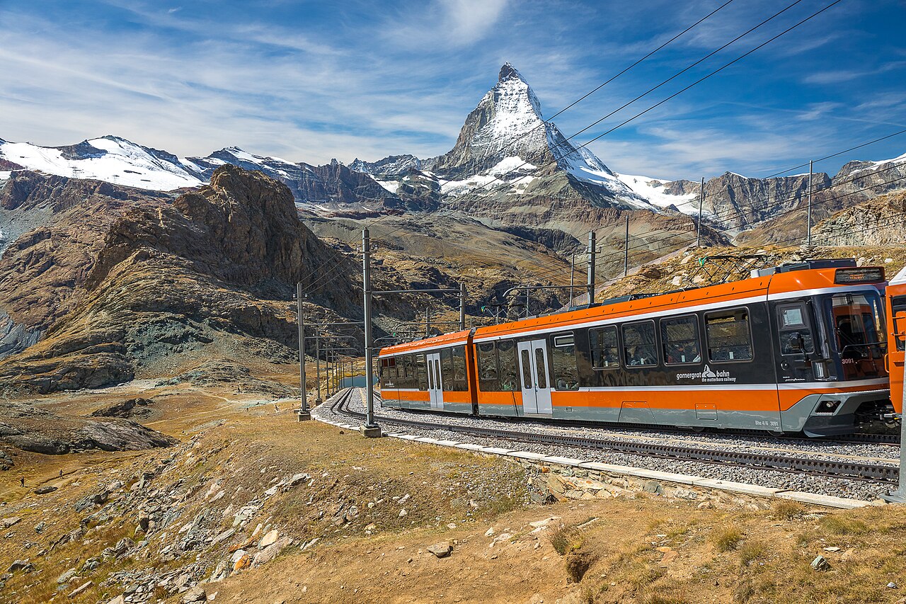 An orange mountain train crosses a grassy slope beneath a sharp pyramid-shaped peak dusted with snow.