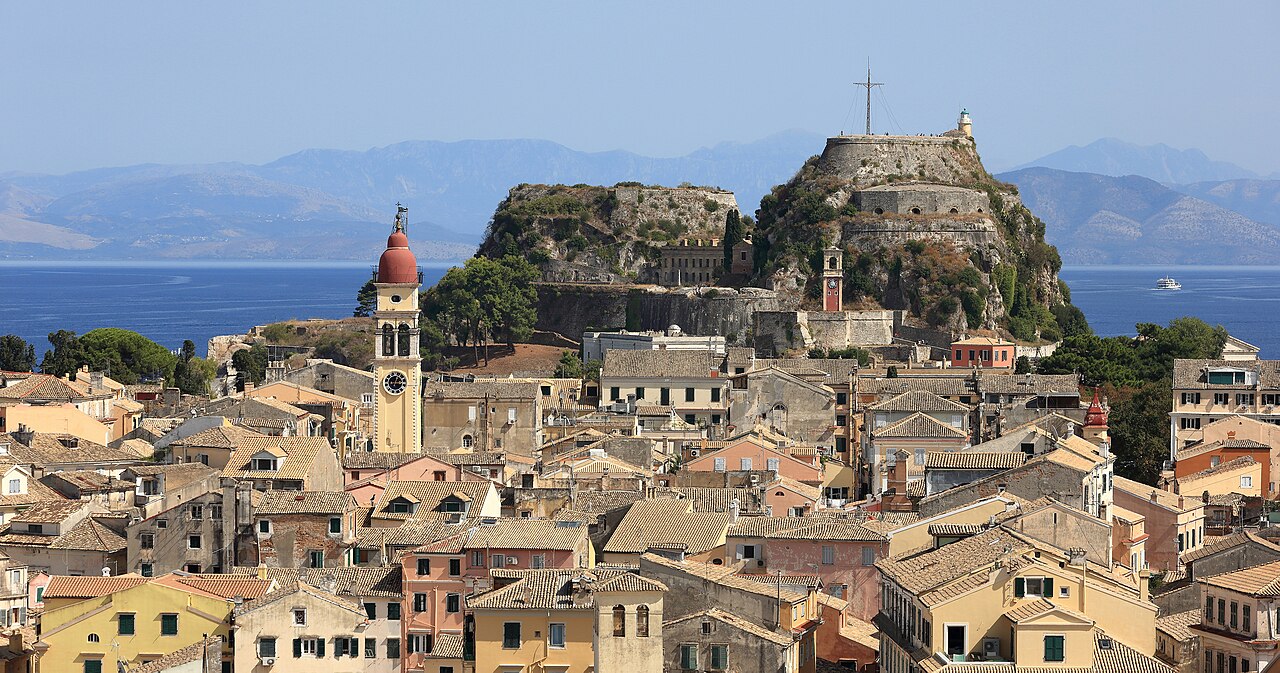 Pale ochre houses and a red-domed bell tower crowd below a rocky fortress hill, with sea and mountains behind.