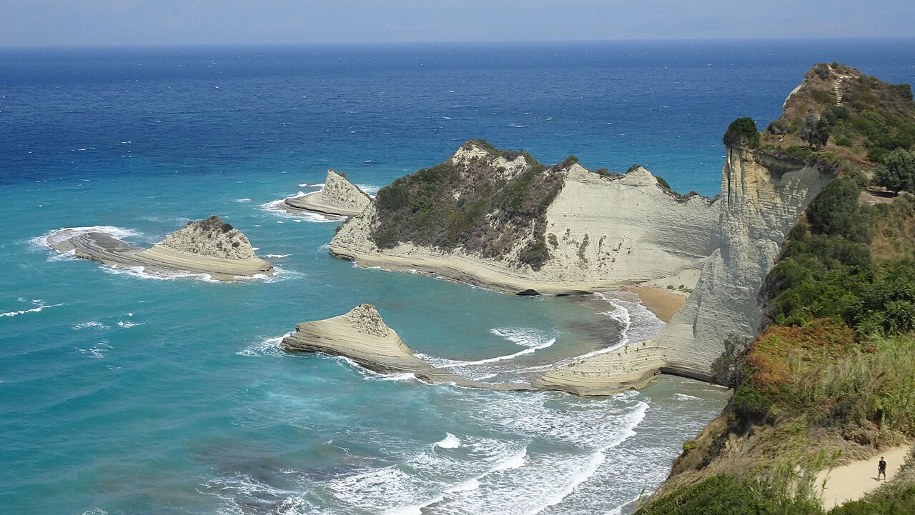 Pale layered cliffs and small rocky islets rise from bright turquoise water with surf breaking on a narrow beach.