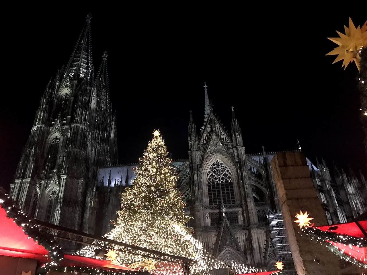 A tall Christmas tree covered in white lights stands among market stalls beneath the dark Gothic towers of a cathedral.