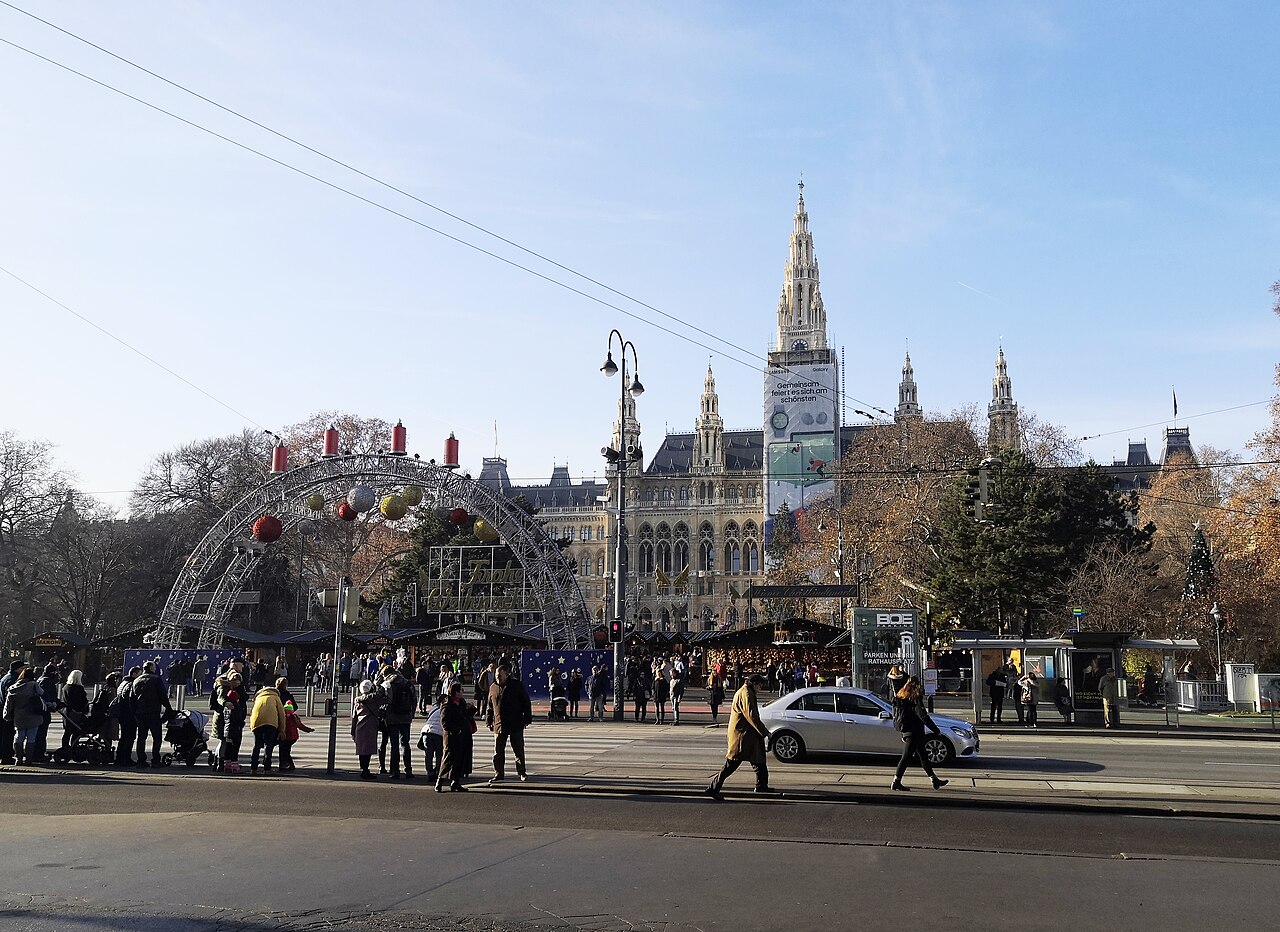 A spired neo-Gothic city hall rises behind a market entrance arch and stalls, with a tram stop in the foreground.