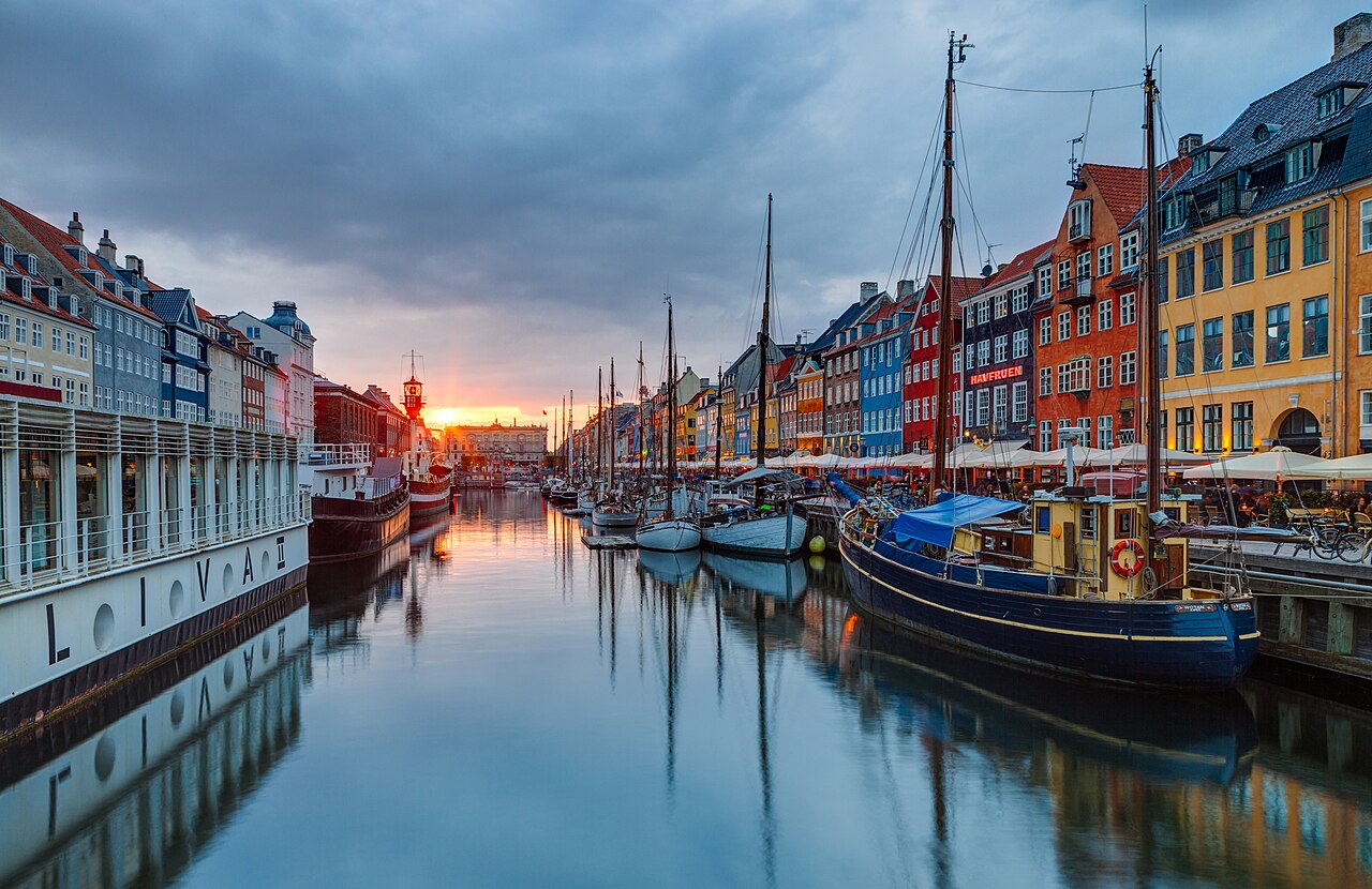 Wooden sailing ships are moored along a still canal lined with tall, brightly painted townhouses under a glowing sky.