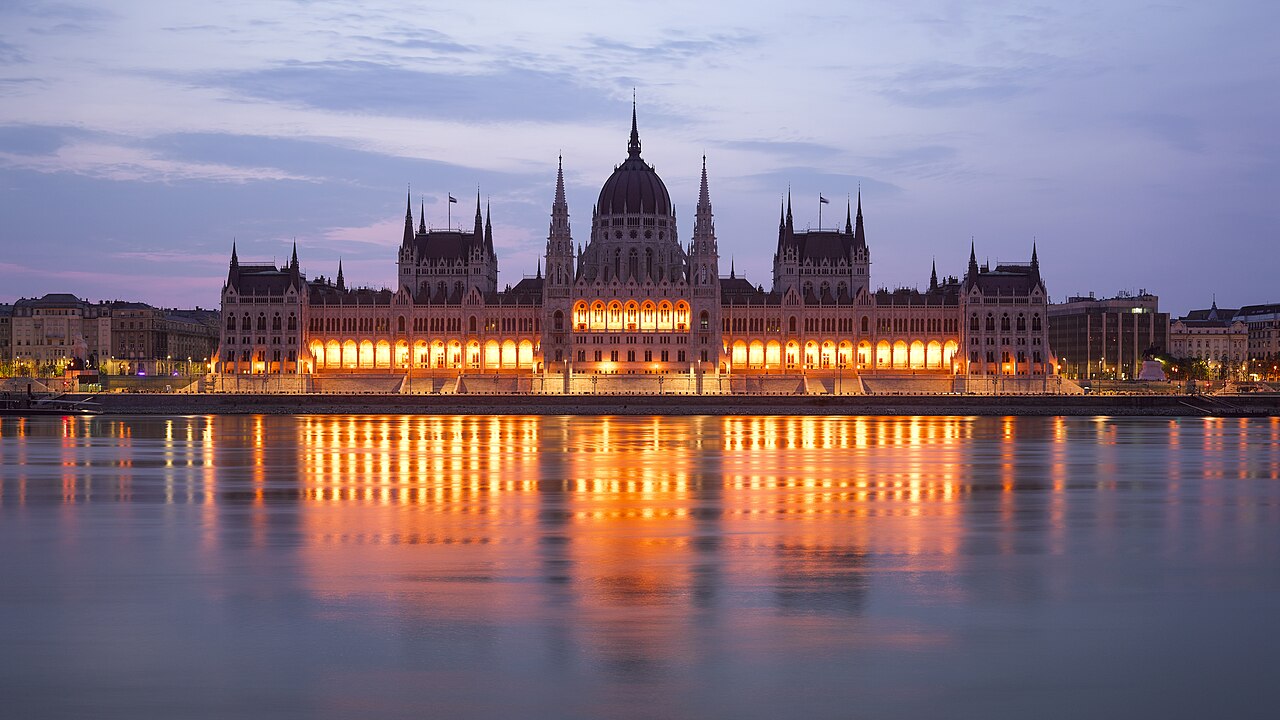A long domed and spired building glows orange along a riverbank, its lights reflected in the calm water.