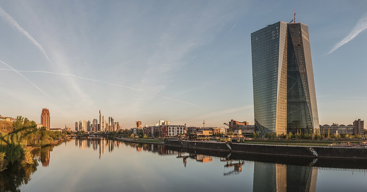 A twisting glass tower stands beside a still river at sunrise, with a cluster of skyscrapers small in the distance.