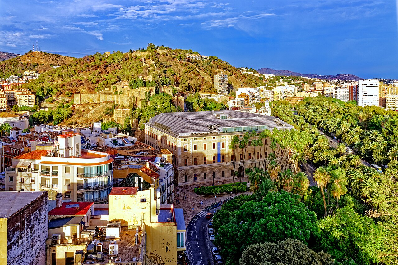City rooftops and a large pale civic building sit below a green hill topped with old fortress walls.