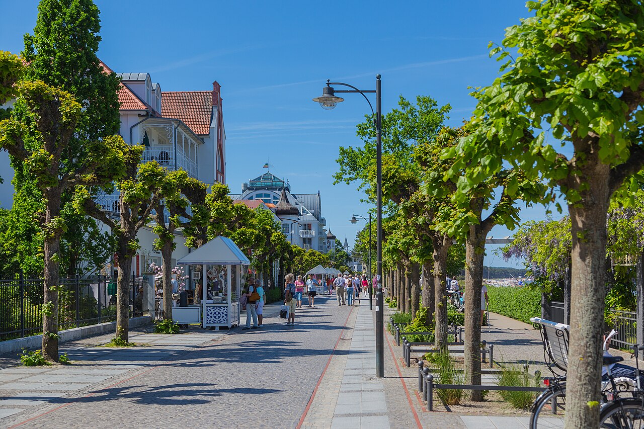 A paved seaside promenade runs between rows of clipped trees past white villas and a small pavilion in bright sun.