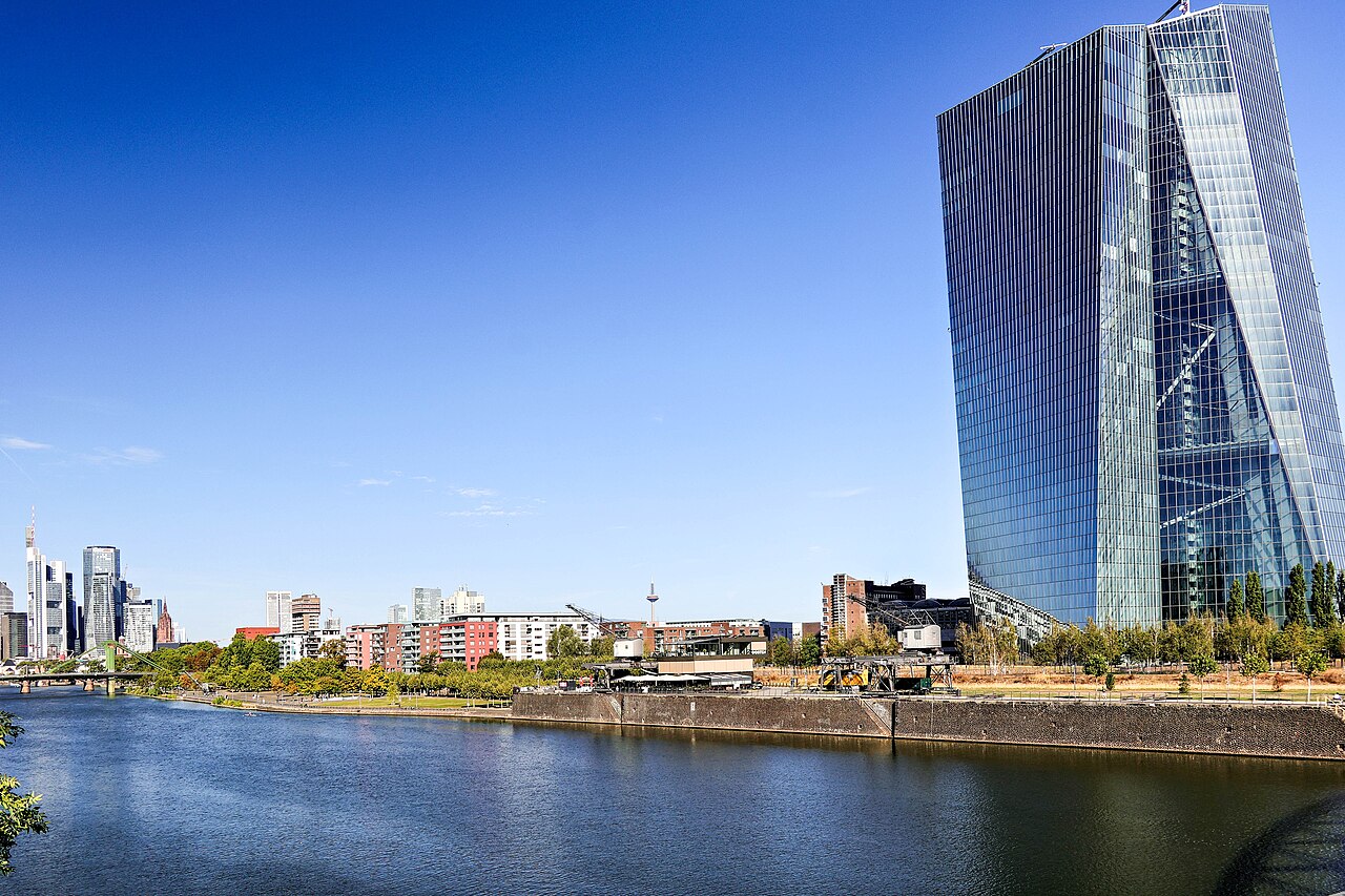 A tall angular glass tower stands on a riverbank under a deep blue sky, with a distant skyline to the left.