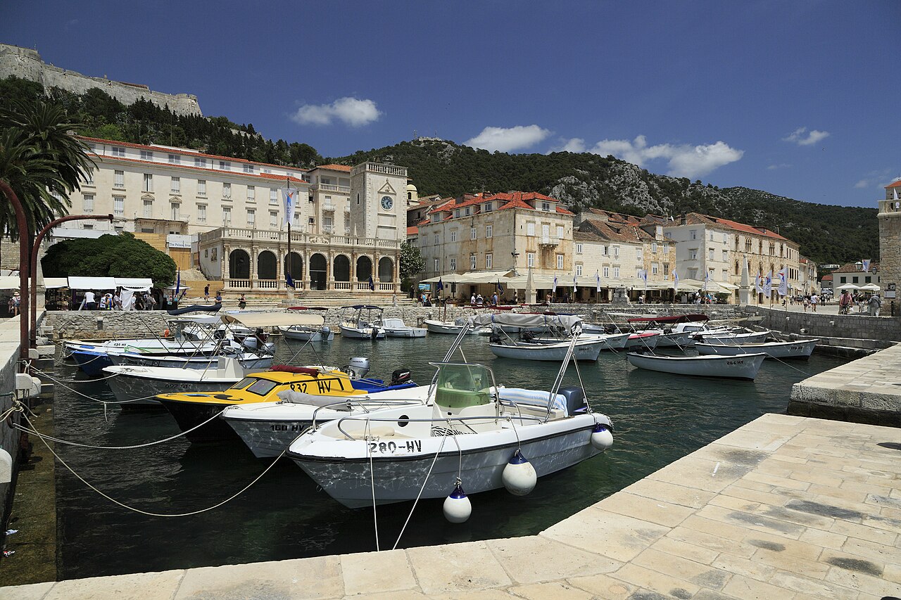 Small motorboats are moored in a stone-walled basin in front of an arcaded pale building and a clock tower.