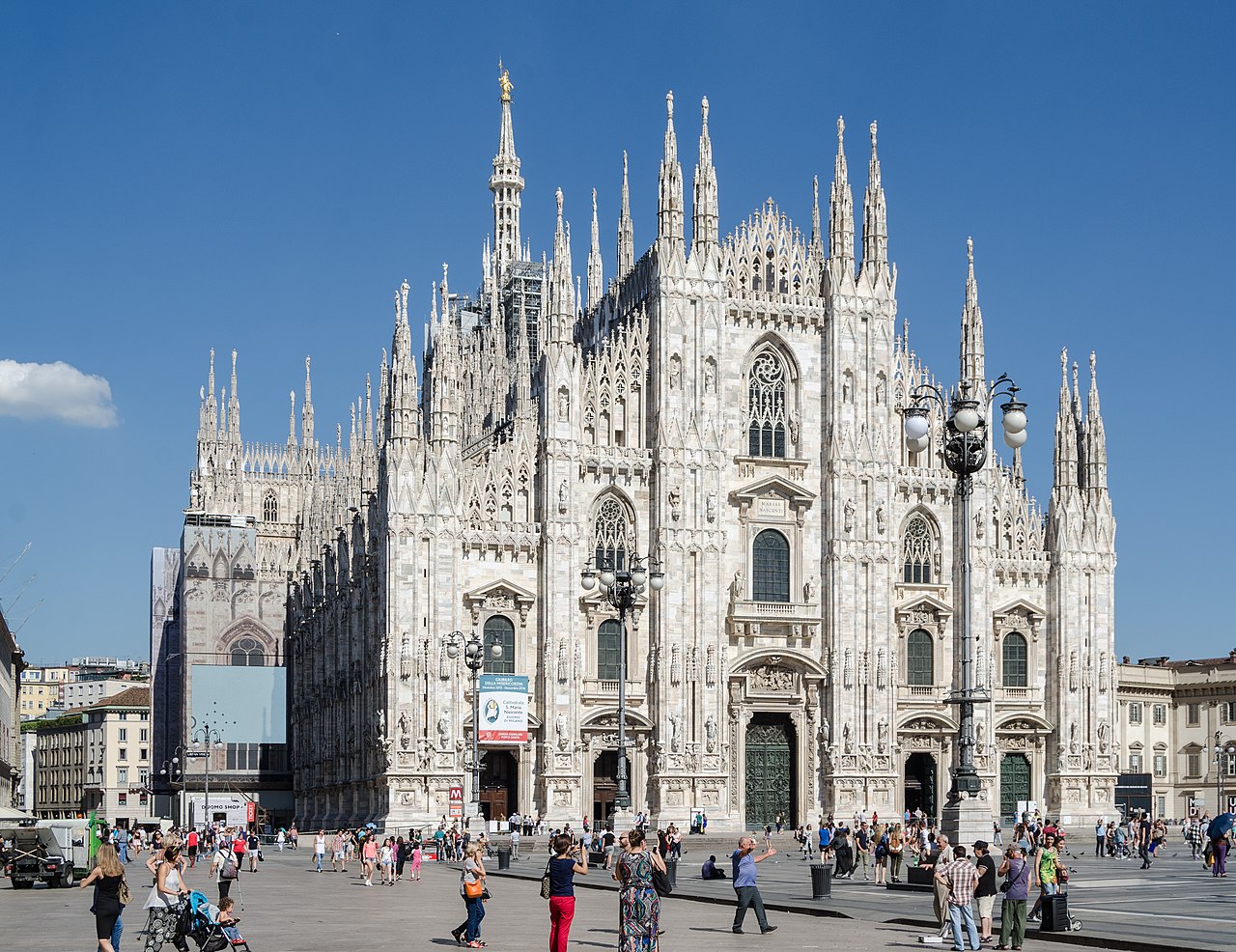 A white marble Gothic cathedral bristling with spires stands over a wide square dotted with small figures.