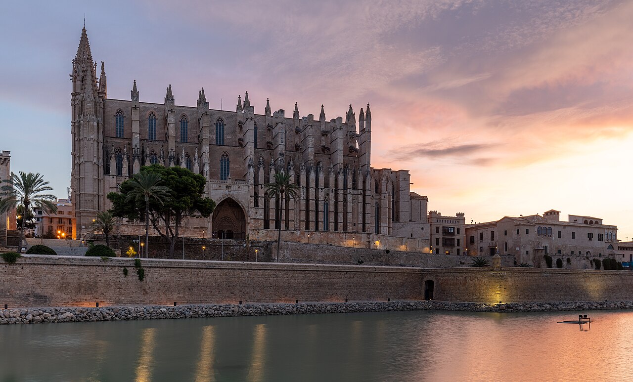 A long Gothic cathedral with rows of buttresses stands above old stone walls and still water under a pink evening sky.