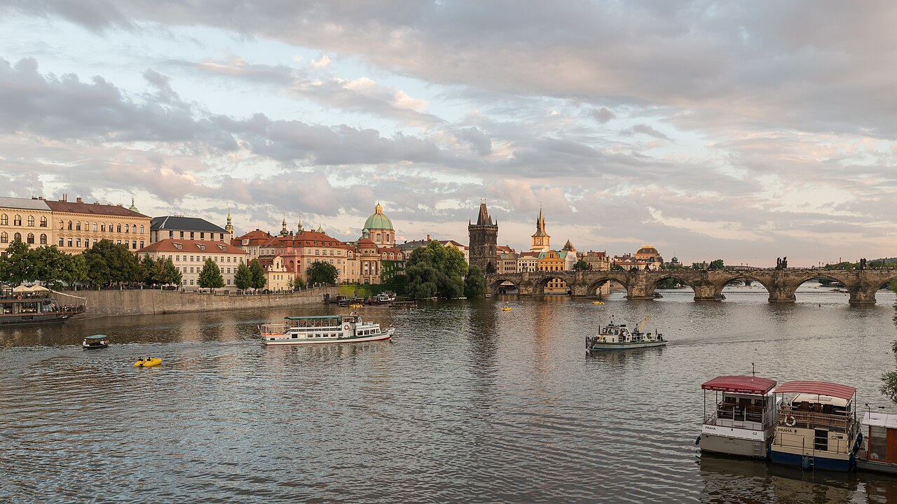 A wide river with tour boats and a long stone arched bridge, domes and towers on the far bank under a cloudy evening sky.