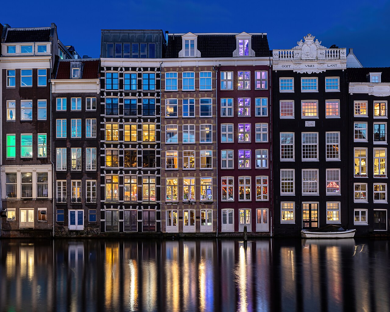 A row of tall narrow houses with lit windows of many colours standing directly on the water, reflected below.