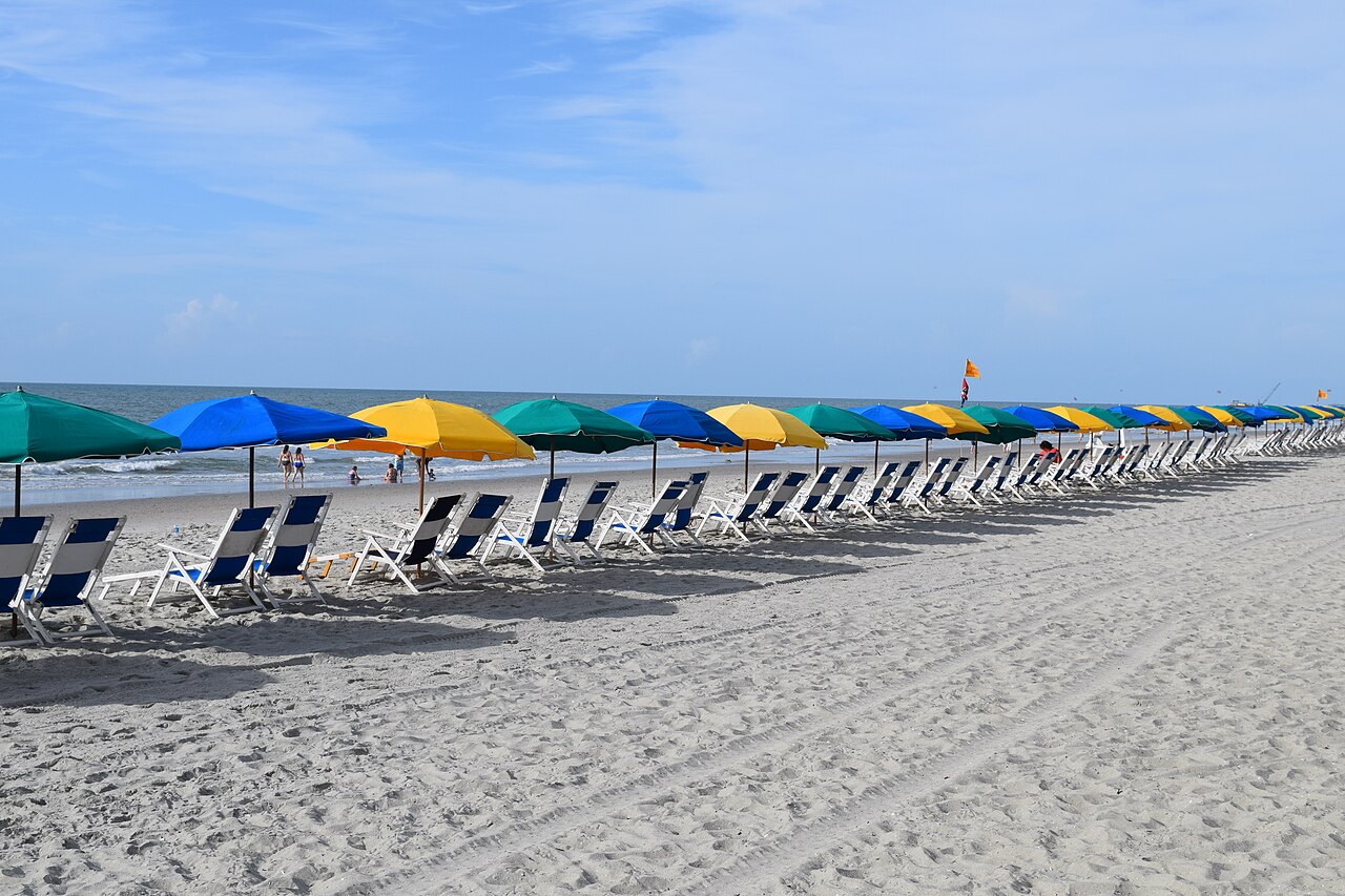 A long line of blue, green and yellow beach umbrellas with empty loungers on pale sand beside the sea.