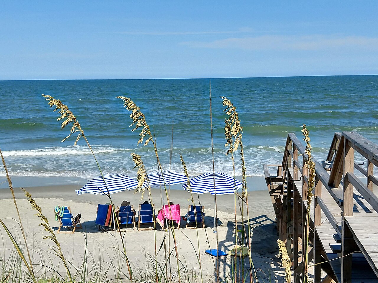 Sea oats in the foreground, a wooden beach walkway and a few umbrellas and chairs at the edge of the surf.
