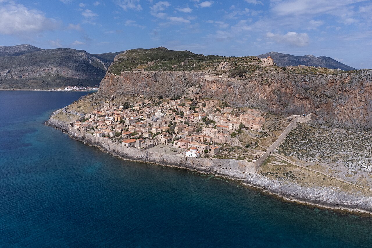 A historic stone town built on the slope of a rocky headland surrounded by blue sea water.