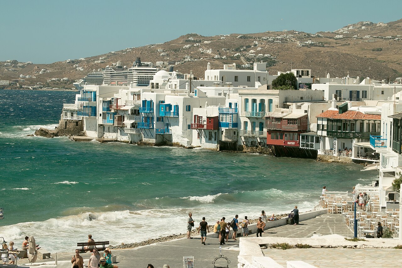 White houses with coloured wooden balconies built at the water's edge, waves breaking below and a bare hill behind.