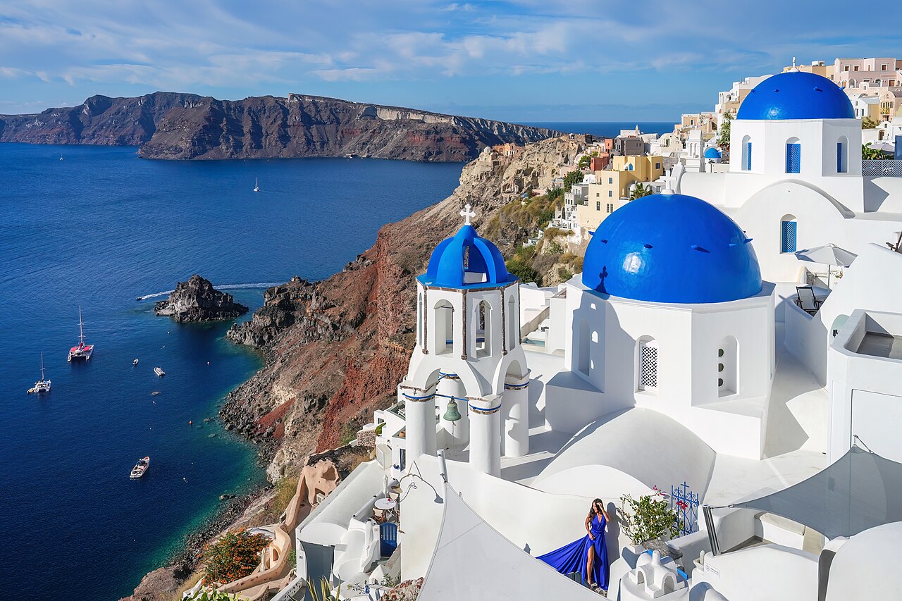 Three blue church domes and a white bell tower on a steep hillside of white houses above a deep blue bay.