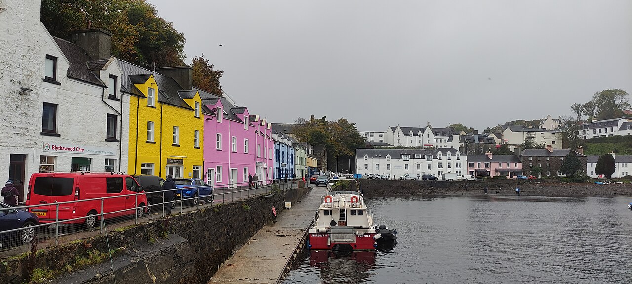 A row of yellow, pink and blue houses along a stone quay, with a small red and white boat moored below.