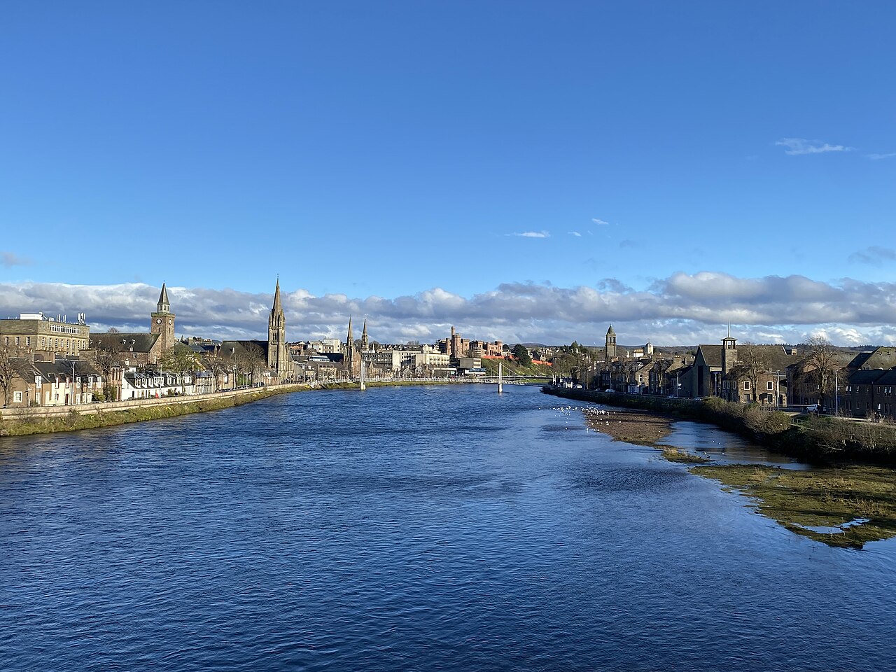 A broad blue river under a clear sky, with church spires and low buildings along both banks.