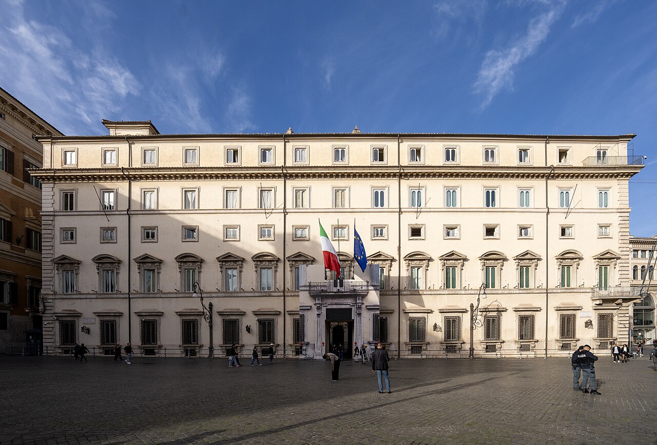 A long pale five-storey palace with flags over its entrance, facing an open paved square.
