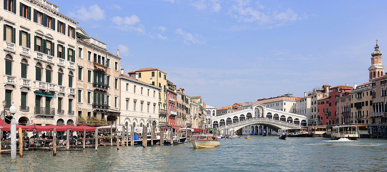 A wide canal lined with palaces and moored boats curving towards a white stone arched bridge.