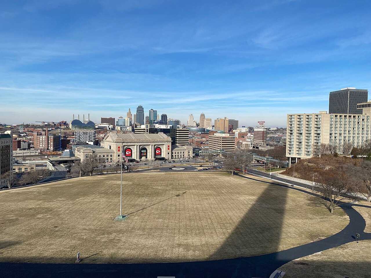 A wide lawn leading to a large stone station building, with downtown towers on the horizon under a blue sky.