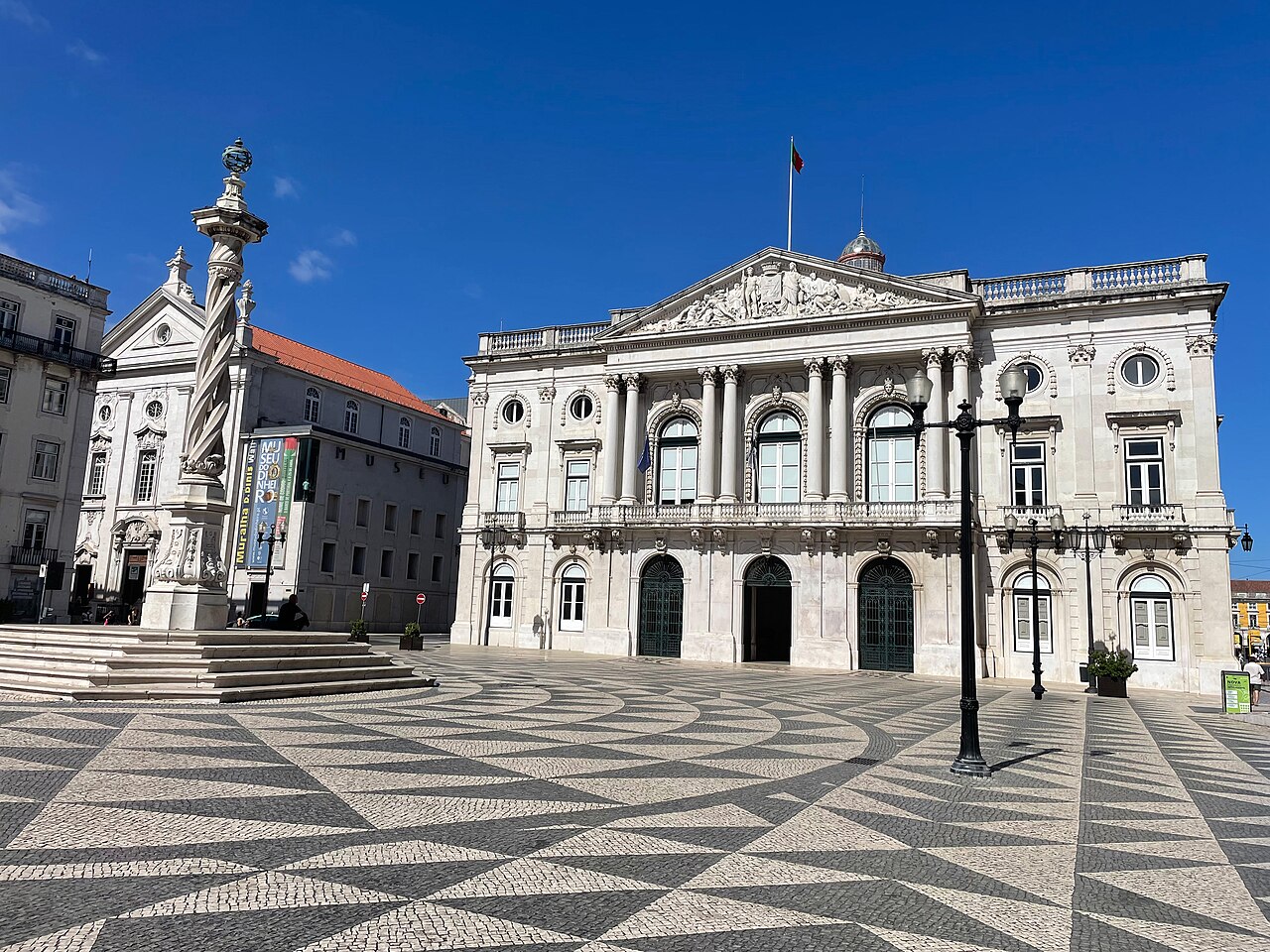 A wide grand square paved in black and white geometric patterns leading to a classical city hall building with columns and a pediment.