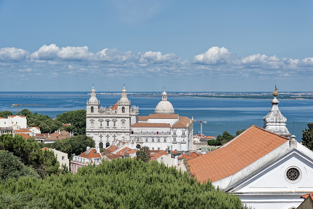 An elevated view over red tile roofs of historical buildings stretching toward a large church and blue water under a partly cloudy sky.