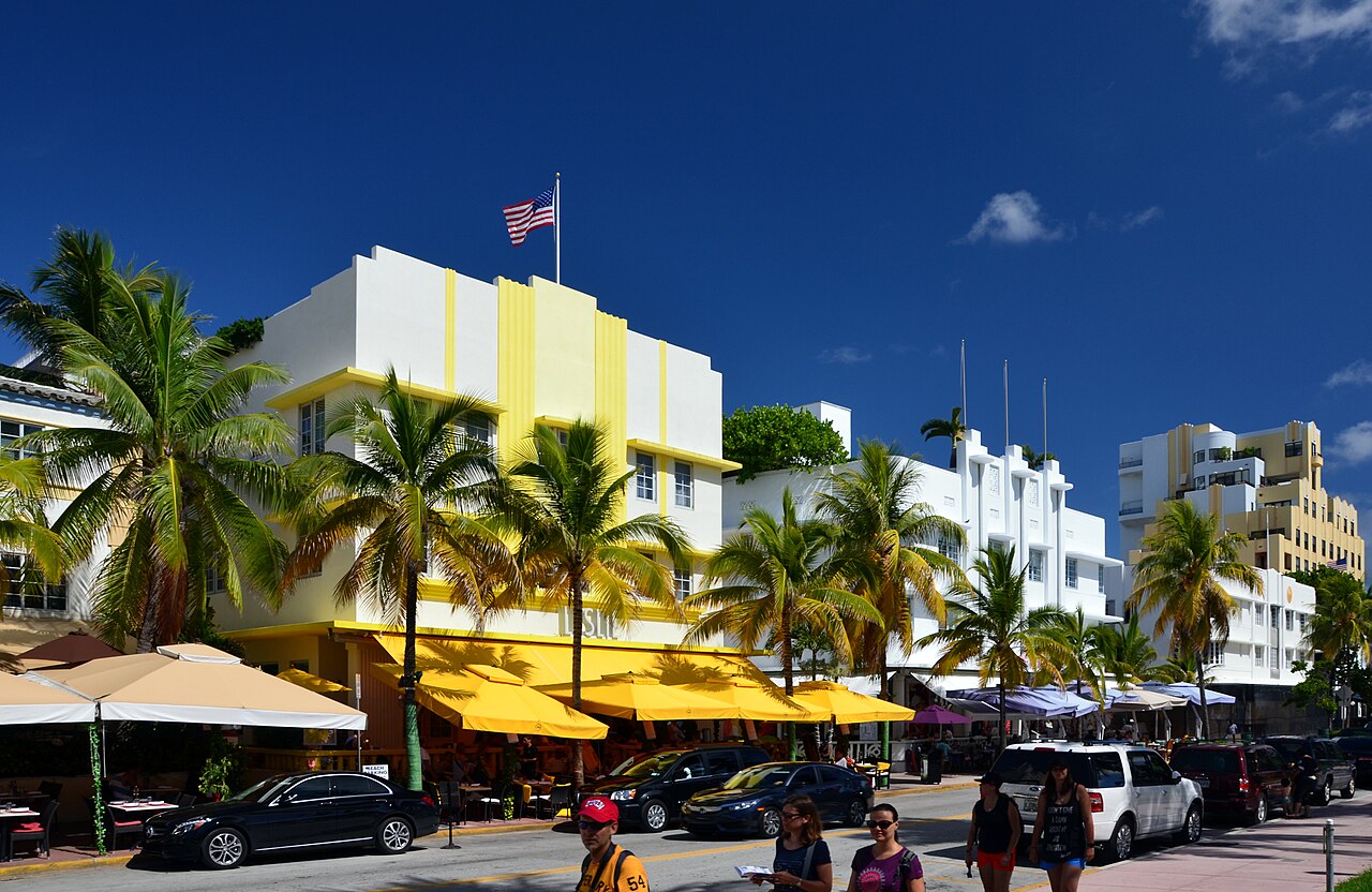 White and yellow Art Deco hotel fronts behind tall palm trees, with cafe umbrellas along the pavement.