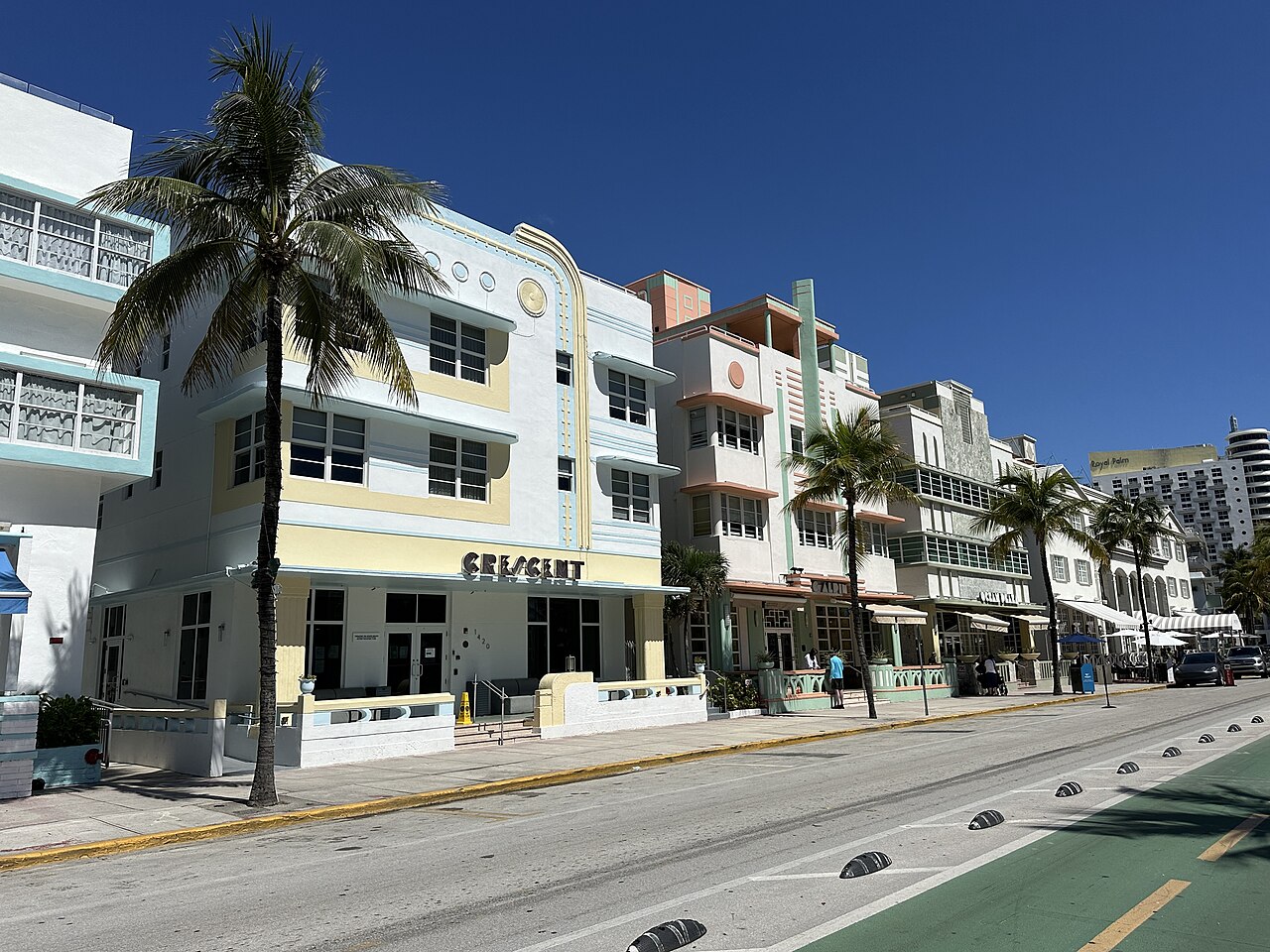 A row of pastel low-rise hotels with palm trees in front, lining a street under a deep blue sky.