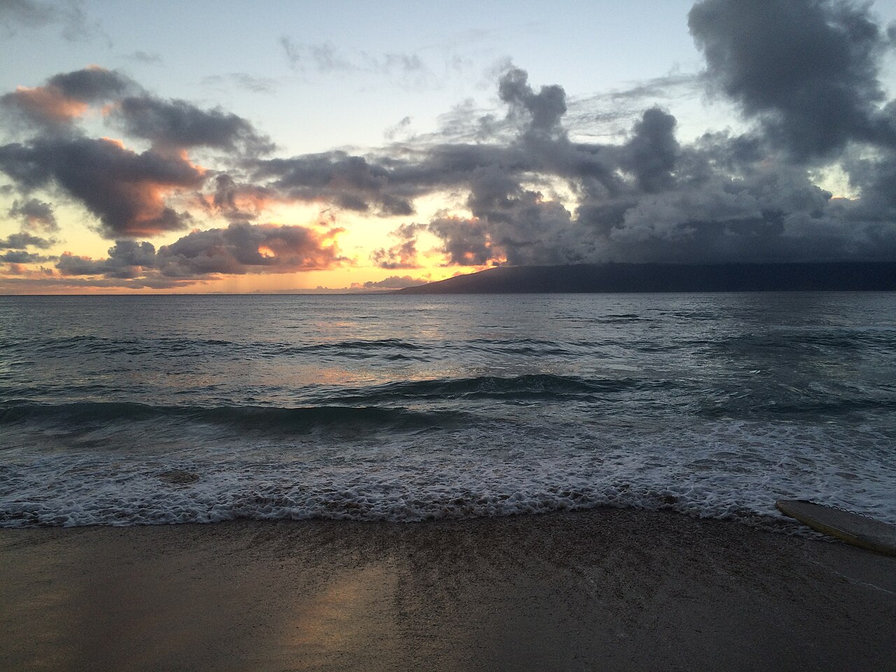 Small waves breaking on dark sand under heavy clouds, with a band of orange light on the horizon.