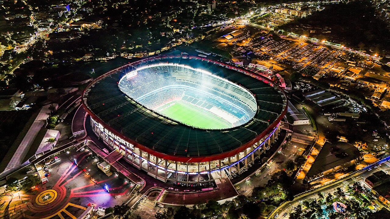 A floodlit oval stadium with a bright green pitch, seen from above and surrounded by city lights at night.