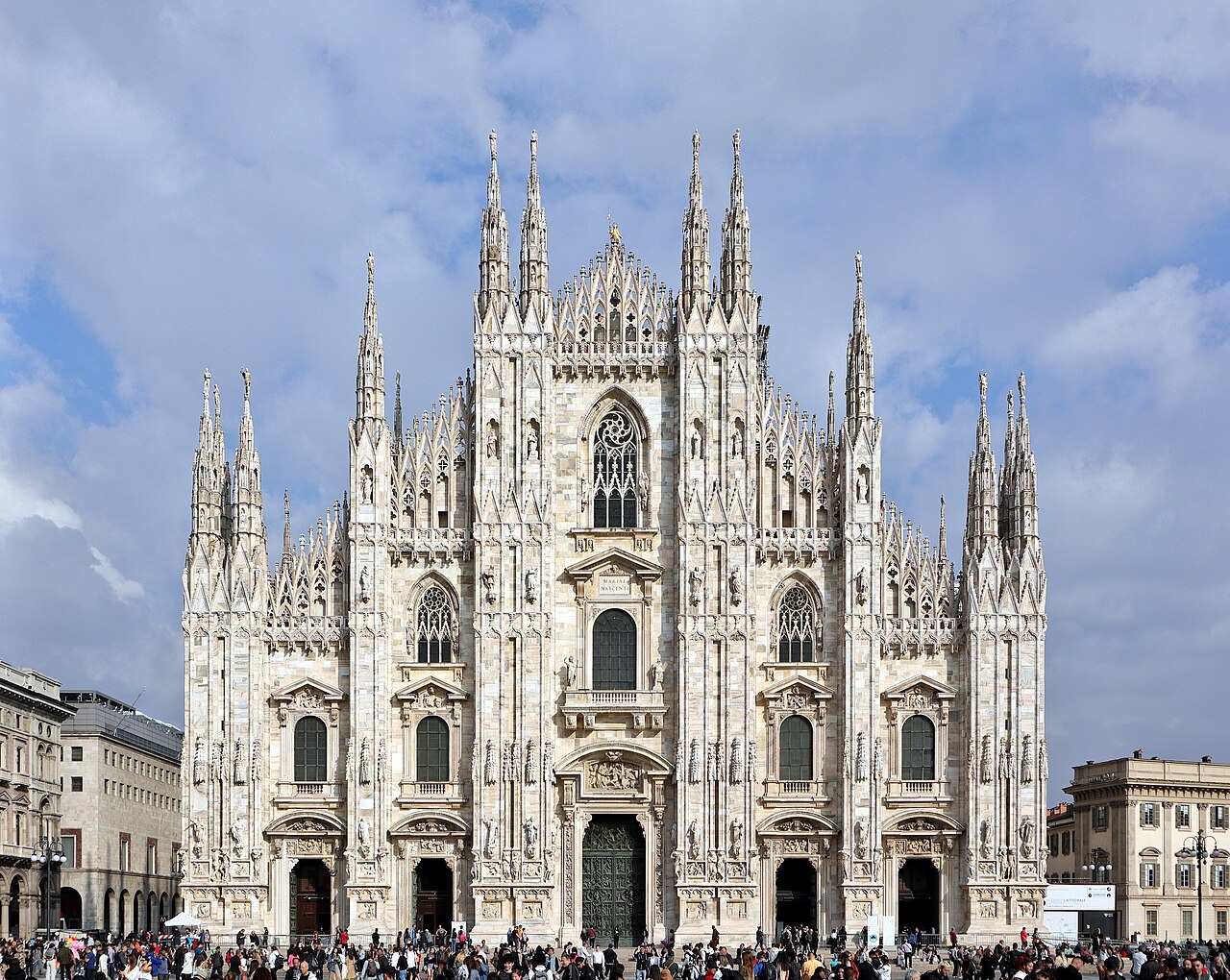 A white marble Gothic cathedral front covered in spires and statues, under a sky of broken cloud.