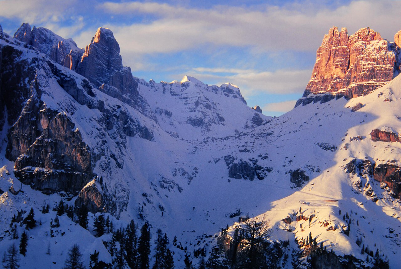 Snowy slopes in blue shadow below jagged rock peaks lit orange by low sun.