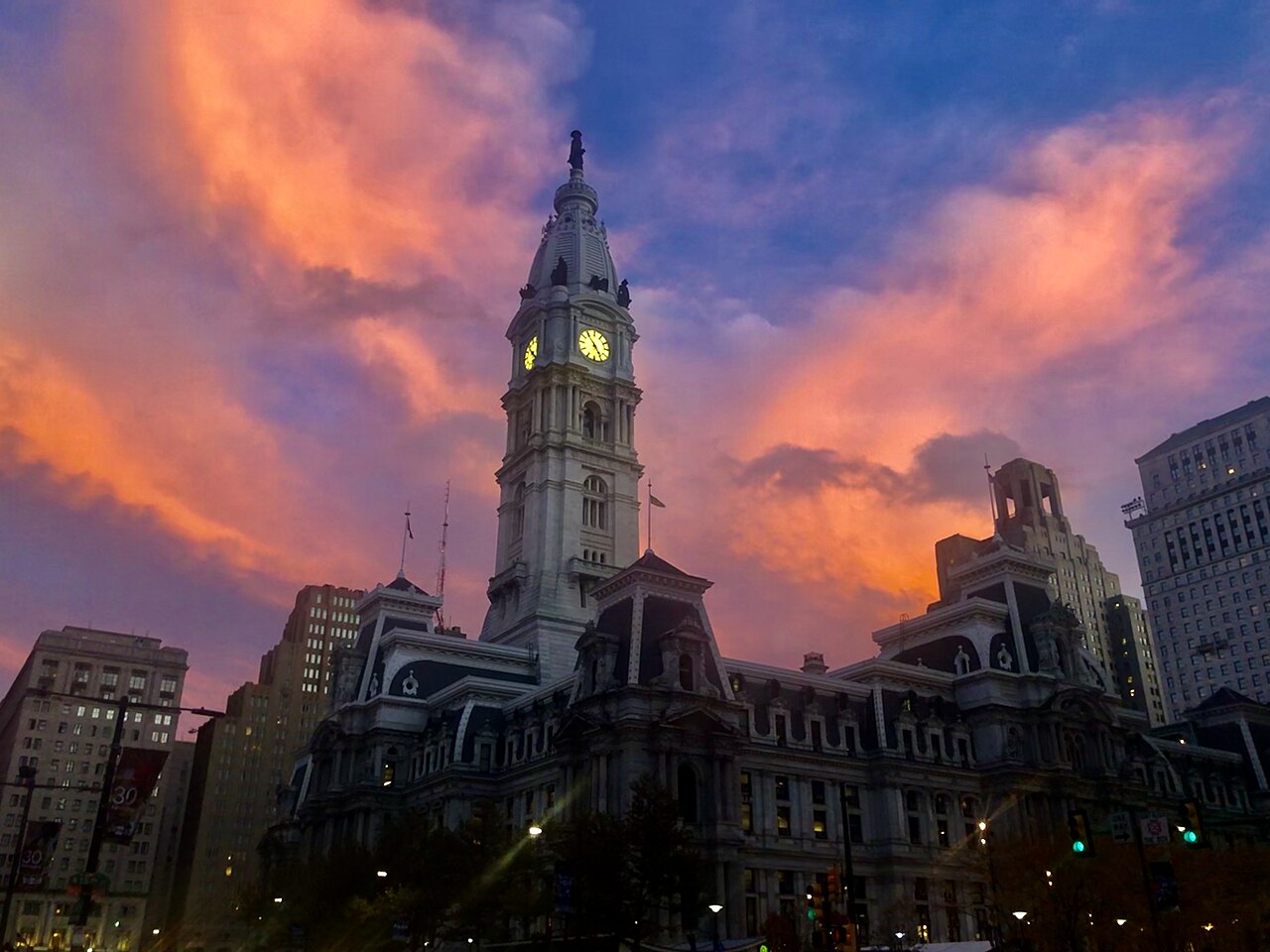 A stone clock tower and mansard roofs in silhouette against orange and purple evening clouds.