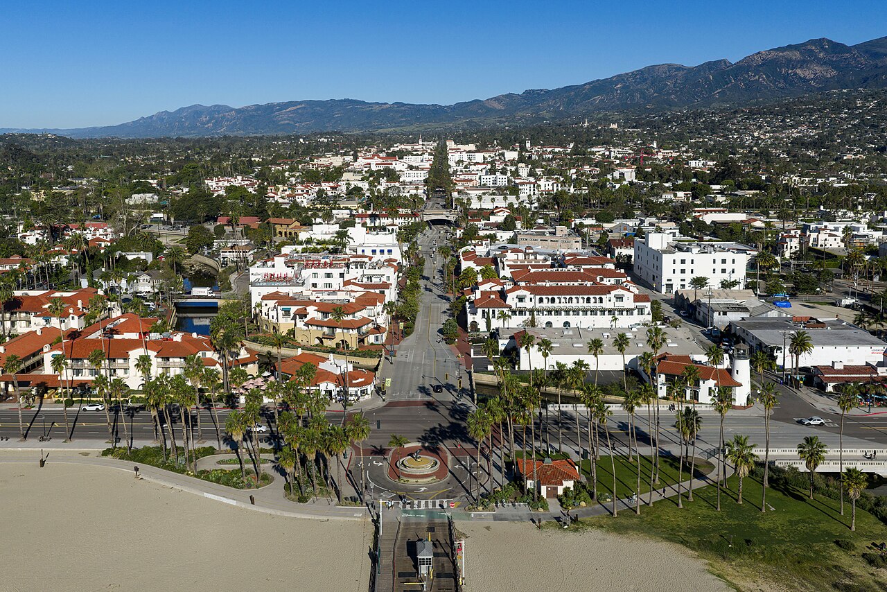 An aerial view up a straight street lined with white buildings and red tile roofs, running from the beach towards mountains.