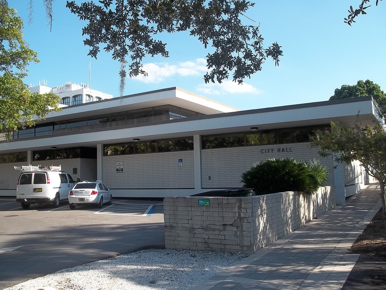 A low-rise modern municipal building made of light brick with wide horizontal windows and a paved driveway in front.
