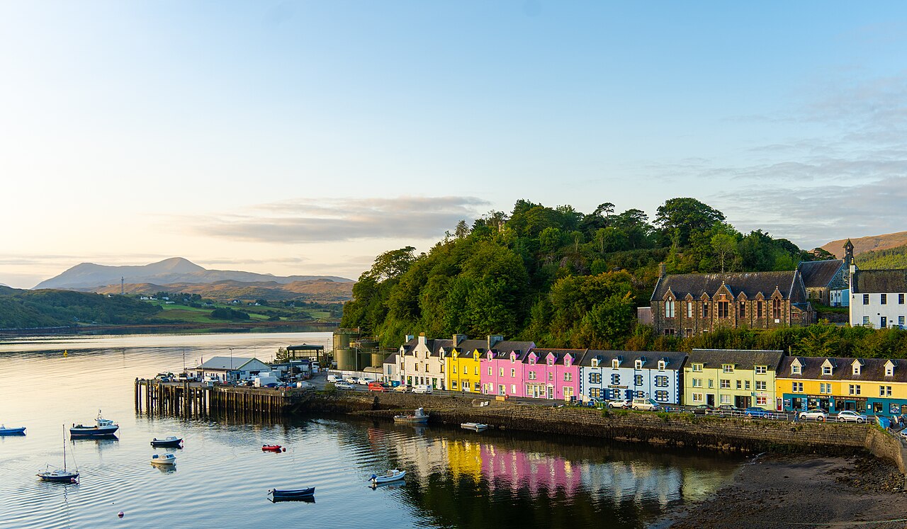A row of pink, blue, yellow and green houses on a quay below a wooded hill, reflected in still water.