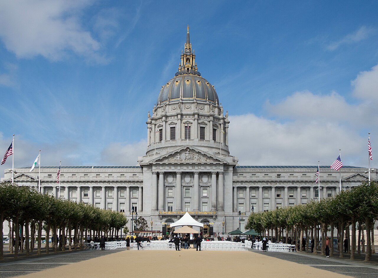 A wide grey stone building with a tall dome, behind rows of pruned trees on a plaza.