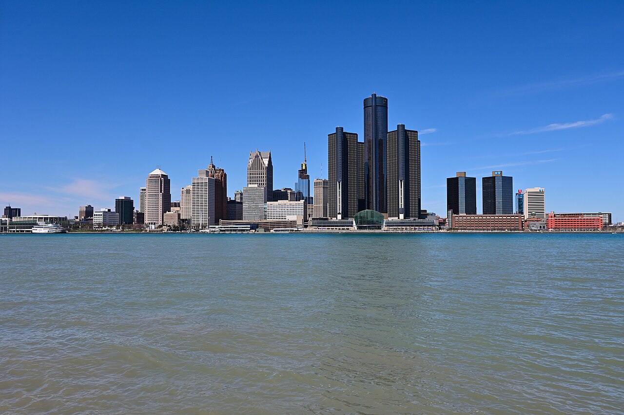 A cluster of dark glass towers and older office buildings rising behind a wide river on a clear day.