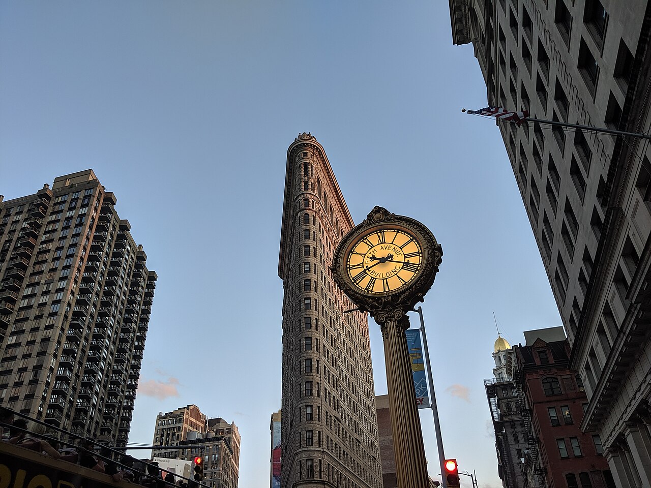 A narrow wedge-shaped stone tower seen from the street at an angle, with a gold-faced street clock in front of it.