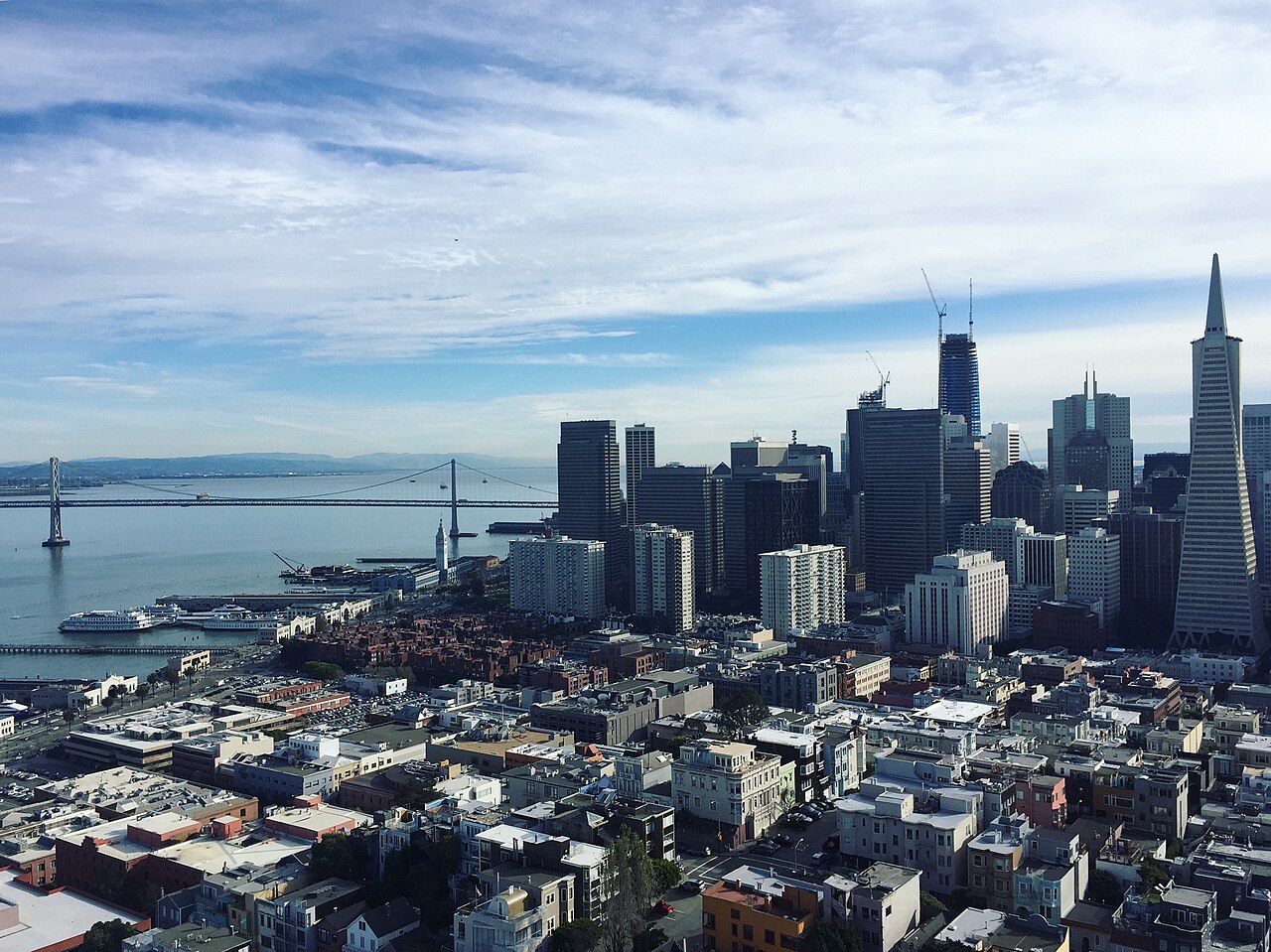 Rooftops of low buildings leading to a group of downtown towers, with a bay and a long bridge to the left.