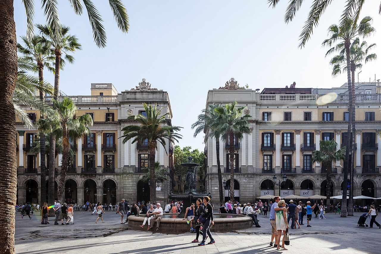 A paved square ringed by pale arcaded buildings and tall palm trees, with a fountain in the middle.