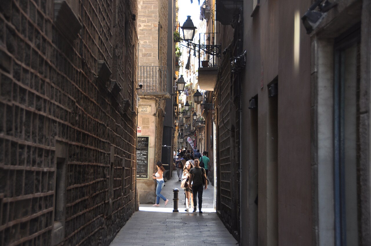 A shaded alley between tall stone walls opening onto a sunlit street with a few walkers in the distance.
