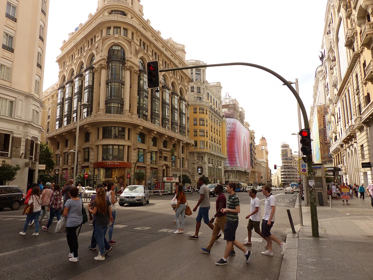 A wide city avenue lined with ornate stone buildings, with people crossing the road in the foreground.