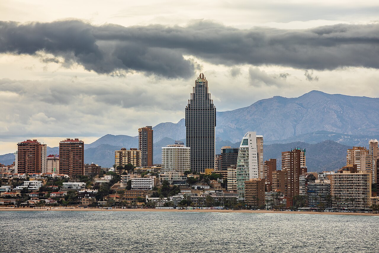 A line of tall apartment and hotel towers along a shore, with mountains and heavy cloud behind them.